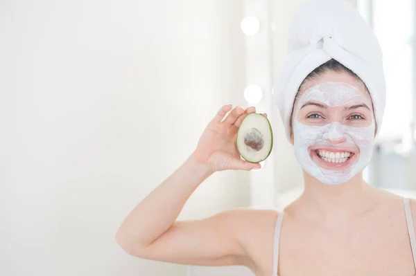 Kobieta z ręcznikiem na włosach i glinianą maską na twarzy trzyma awokado. Dbanie o piękno w domu — Zdjęcie stockowe