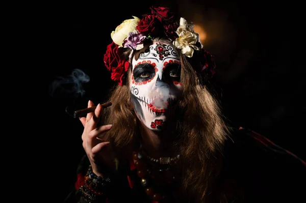 Mulher em Santa Muerte maquiagem em um fundo preto. Halloween menina fumar um charuto vestido como uma morte santa mexicana tradicional. — Fotografia de Stock