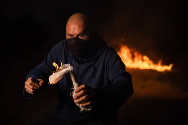 Maskeli bir adam yanan bir şişe tutuyor. Molotof kokteyli.