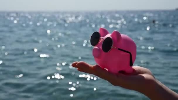 Kobieta trzyma skarbonkę w okularach przeciwsłonecznych na tle morza. Urlop budżetowy. — Wideo stockowe