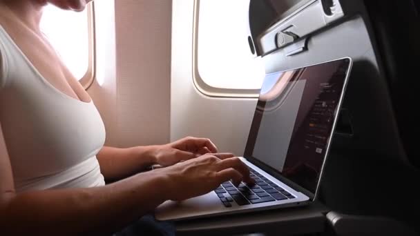 Mulher sem rosto em uma máscara funciona em um laptop em um avião enquanto sentado à janela durante o voo. Freelancer em uma viagem. — Vídeo de Stock