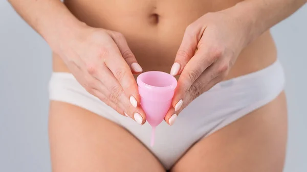 Close-up seorang wanita dengan celana dalam katun putih memegang cangkir menstruasi merah muda dengan latar belakang putih. Alternatif untuk pembalut dan bantalan — Stok Foto