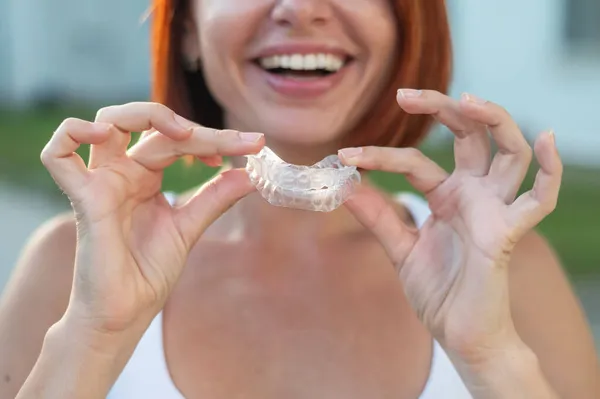 Red-haired Caucasian woman holding transparent mouthguards for bite correction outdoors. A girl with a beautiful snow-white smile uses silicone braces — Stock Photo, Image