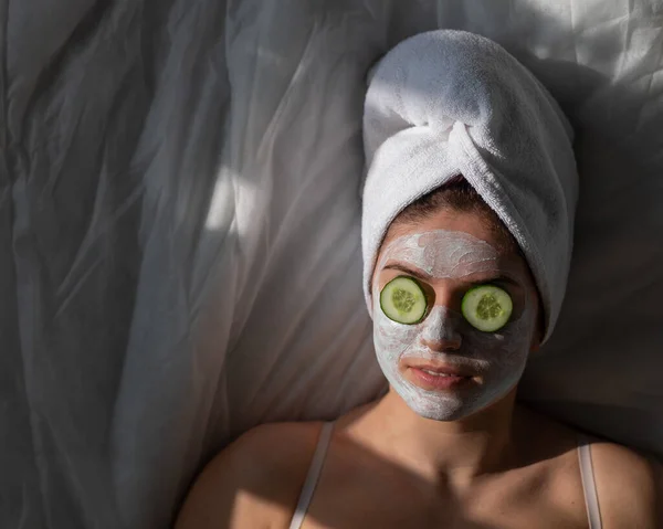 Una mujer con una toalla en el pelo y en una mascarilla de barro y pepinos frente a sus ojos yace sobre la sábana — Foto de Stock