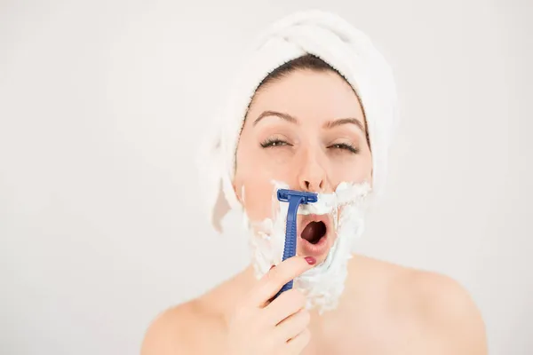 Femme caucasienne joyeuse avec une serviette sur la tête et mousse à raser sur son visage tient un rasoir sur un fond blanc — Photo