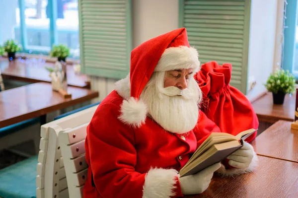 Ο Άγιος Βασίλης κάθεται σε ένα καφέ και διαβάζει ένα βιβλίο. — Φωτογραφία Αρχείου