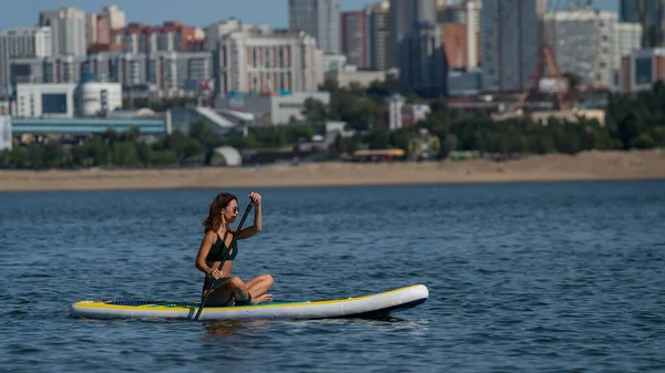 Mujer caucásica está montando una tabla de SUP en el río en la ciudad. Deportes de verano. — Foto de Stock