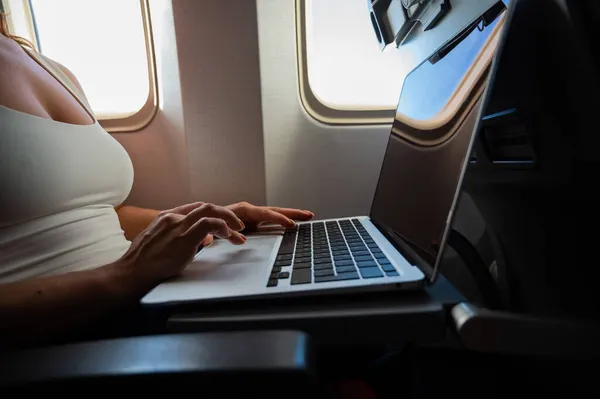 La mujer sin rostro en una máscara trabaja en un portátil en un avión mientras está sentada junto a la ventana durante el vuelo. Freelancer en un viaje. — Foto de Stock