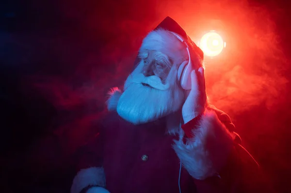 De kerstman luistert naar muziek met koptelefoon op een feestje. Blauw rood neon licht en rook. — Stockfoto