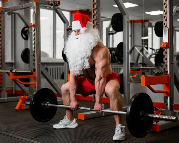 Święty Mikołaj na siłowni. Muskularny mężczyzna z nagim tułowiem ćwiczący z hantlami. — Zdjęcie stockowe