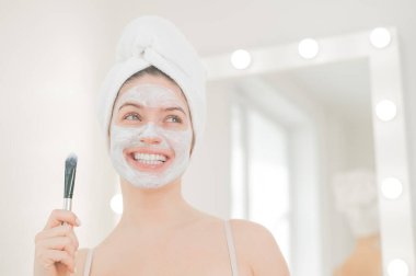 Saçında havlu olan güzel beyaz kadın kilden bir maske takıyor. Evdeki güzellikle ilgilenmek.