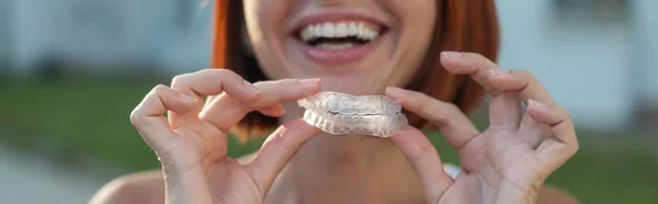 Mulher caucasiana de cabelos vermelhos segurando protetores bucais transparentes para correção de mordida ao ar livre. Uma menina com um belo sorriso branco-neve usa aparelho de silicone — Fotografia de Stock