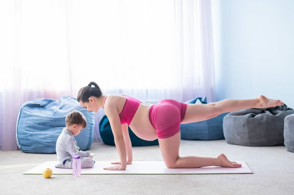 A pregnant woman in sportswear does gymnastics while her son plays in the mobile. Girl on yoga with her child. Fitness mom and baby. — Stock Photo, Image
