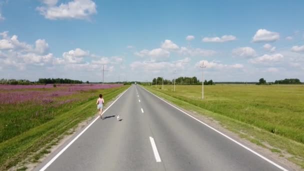 Una donna caucasica corre lungo l'autostrada con un cane. Campi di fireweed. — Video Stock