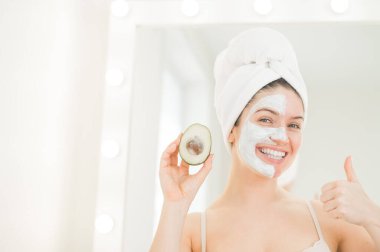 Saçında havlu ve kilden maskeli bir kadın bir avokado tutuyor. Evdeki güzellikle ilgilenmek.