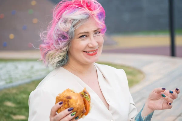 Vit kvinna med lockigt färgat hår som äter burgare. Dåliga matvanor och kärlek till snabbmat — Stockfoto