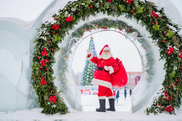 Jultomten poserar med en påse med gåvor på bakgrunden av juldekorationer utomhus — Stockfoto