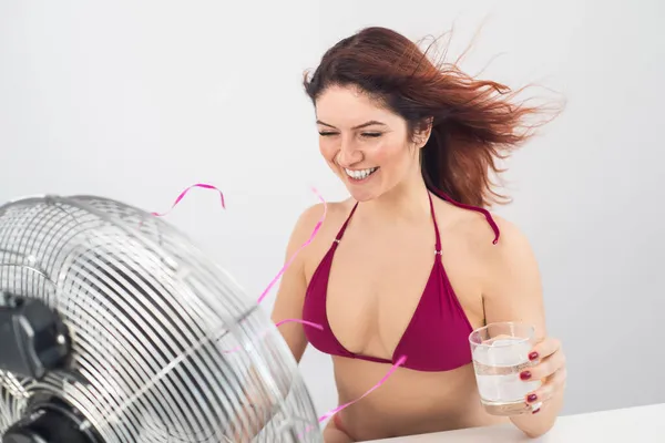 Κοκκινομάλλα χαμογελαστή γυναίκα σε ένα μπικίνι πίνει ένα κρύο ποτό και απολαμβάνει τον άνεμο που φυσάει από έναν ηλεκτρικό ανεμιστήρα σε λευκό φόντο. τον έλεγχο του κλίματος σε μια ζεστή μέρα του καλοκαιριού — Φωτογραφία Αρχείου
