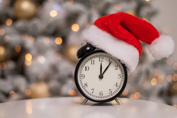 Relógio de alarme em um chapéu de Papai Noel contra o fundo de uma árvore de Natal. — Fotografia de Stock