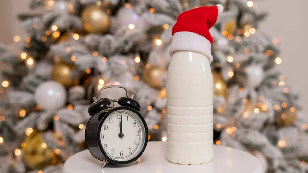 Una botella de leche con un sombrero de Santa Claus y un despertador en el fondo de un árbol de Navidad. Golosinas para Santa. — Foto de Stock
