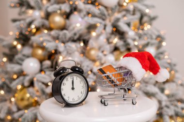 Noel hediyeleri alışveriş zamanı. İçinde kredi kartı olan Noel Baba şapkalı bir alışveriş arabası ve Noel ağacının yanında bir çalar saat..