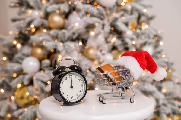 Χριστουγεννιάτικα δώρα για ψώνια. Ένα τρόλεϊ με καπέλο Σάντα με πιστωτική κάρτα μέσα και ξυπνητήρι δίπλα στο χριστουγεννιάτικο δέντρο.. — Φωτογραφία Αρχείου