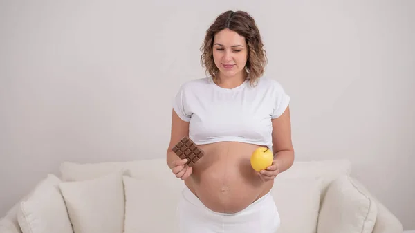 Uma grávida está segurando uma maçã e uma barra de chocolate. O nono mês de gravidez. — Fotografia de Stock