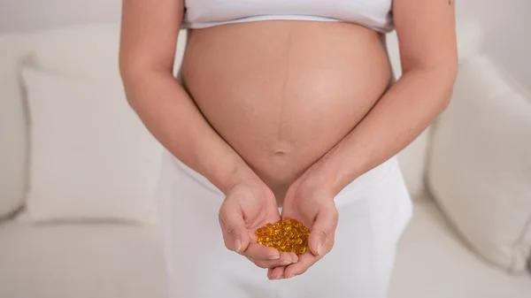 Těhotná žena s nahým břichem drží plnou hrst vitamínů. Průhledné zlaté pilulky. Vitamin D pro nastávající matku. — Stock fotografie