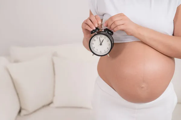 Uma mulher grávida com uma barriga nua segura um despertador. Data prevista de nascimento. — Fotografia de Stock