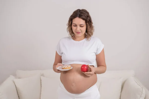 Беременная женщина держит красное яблоко и пончик. Выбор продуктов питания. — стоковое фото
