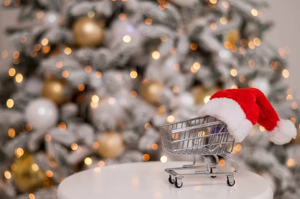 Presentes de Natal tempo de compras. Carrinho de compras em um chapéu de Papai Noel perto da árvore de Natal. — Fotografia de Stock