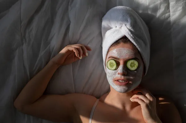 Una mujer con una toalla en el pelo y en una mascarilla de barro y pepinos frente a sus ojos yace sobre la sábana — Foto de Stock