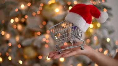 Noel hediyeleri alışveriş zamanı. Noel Baba şapkasıyla Noel ağacının yanında alışveriş arabası tutan kadın..