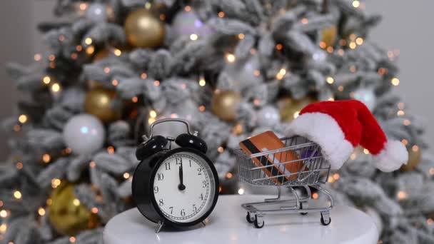 Dags att handla julklappar. En kundvagn med tomteluva med kreditkort och väckarklocka vid granen. — Stockvideo