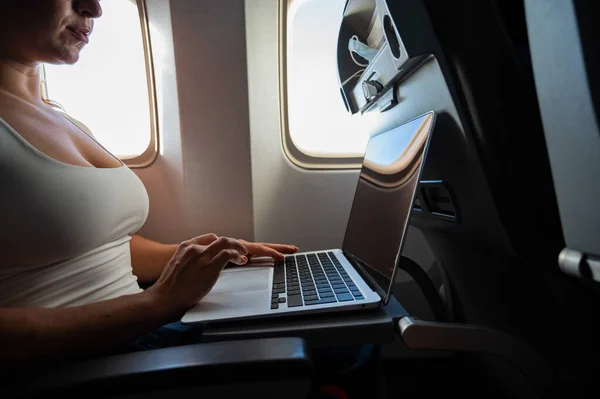 La mujer sin rostro en una máscara trabaja en un portátil en un avión mientras está sentada junto a la ventana durante el vuelo. Freelancer en un viaje. — Foto de Stock