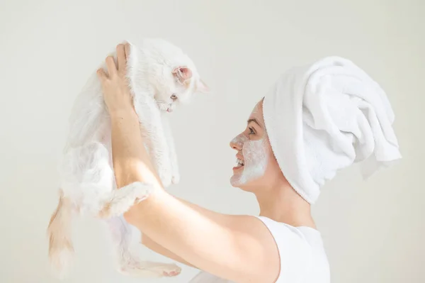 Kobieta z ręcznikiem na włosach i glinianą maską na twarzy trzyma białego puszystego kota na białym tle. Przestrzeń kopiowania — Zdjęcie stockowe