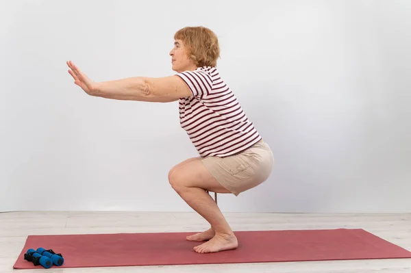 Äldre kvinna gör knäböj på en vit bakgrund. Den gamla damen gör övningar för sin hälsa — Stockfoto