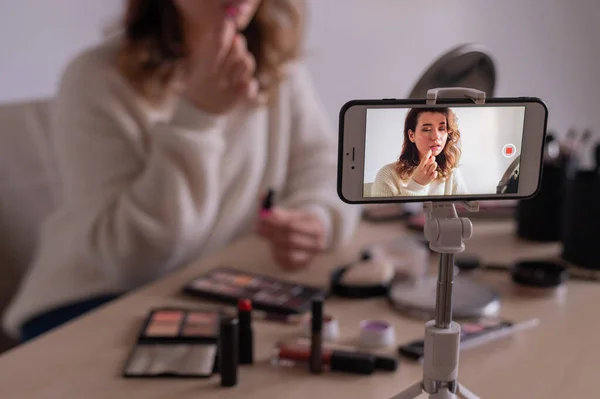 Young caucasian woman is recording an online make-up training video on her mobile. Video blogger girl applies makeup.