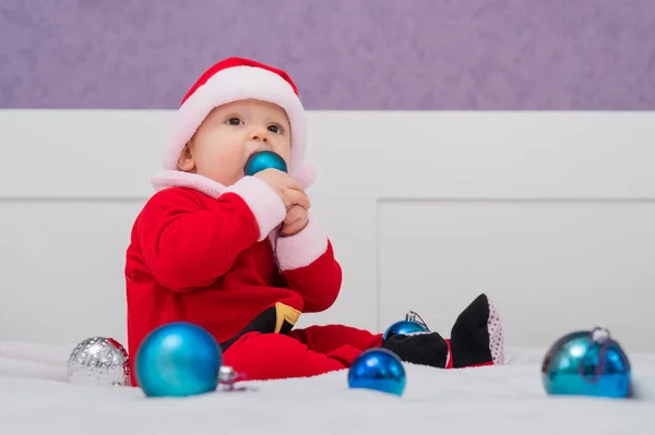 Chłopiec przebrany za Mikołaja bawi się świątecznymi dekoracjami na łóżku. — Zdjęcie stockowe