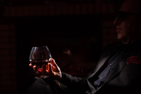 Egy felnőtt férfi poharakkal pálinkát iszik és szivarozik, miközben egy karosszékben ül a kandalló mellett a sötétben. Az elit úriemberek klubjának koncepciója — Stock Fotó