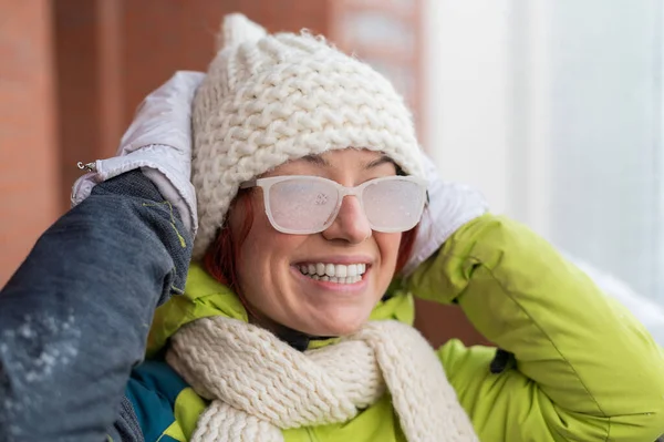 Улыбающаяся белая женщина зимой стоит на балконе кирпичного дома. Счастливая девушка в покрытых льдом очках на улице — стоковое фото