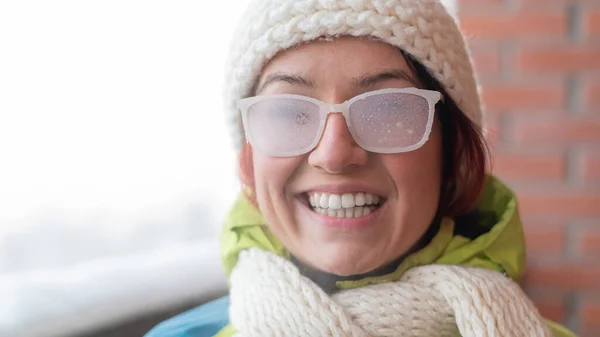 Mulher branca sorridente fica na varanda de uma casa de tijolos no inverno. Menina feliz em óculos cobertos de gelo na rua ao ar livre — Fotografia de Stock