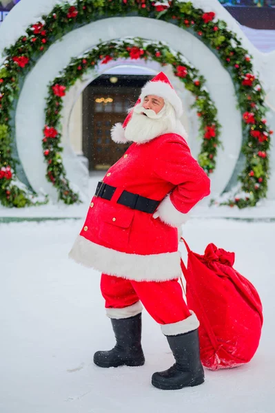 Père Noël posant avec un sac de cadeaux sur le fond des décorations de Noël en plein air — Photo