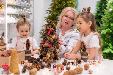 Yaşlı beyaz kadın iki torunuyla Noel için çam kozalağı süslemesi yapıyor.
