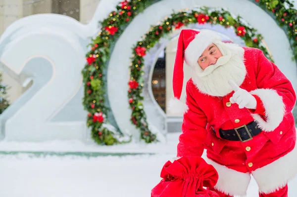 Санта-Клаус позирует с мешком подарков на фоне рождественских украшений на открытом воздухе — стоковое фото