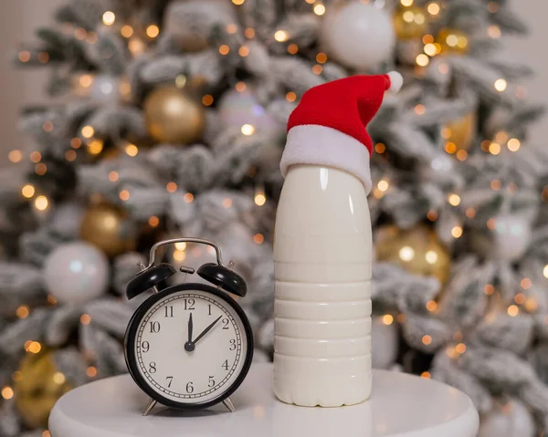 Uma garrafa de leite com um chapéu de Pai Natal e um despertador no fundo de uma árvore de Natal. Doces para Santa. — Fotografia de Stock