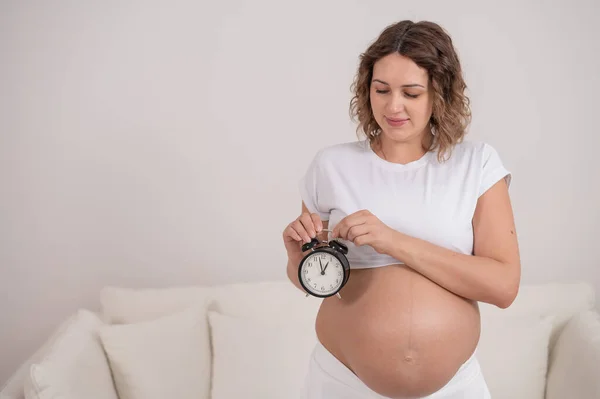 Een zwangere vrouw met een naakte buik houdt een wekker vast. Beoogde geboortedatum. — Stockfoto