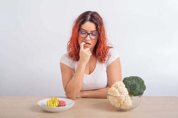 Běloška na dietě sní o rychlém občerstvení. Redhead dívka si vybere mezi brokolicí a koblihy na bílém pozadí. — Stock fotografie