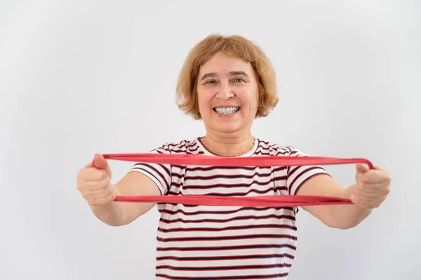 En äldre kvinna är engagerad i fitness med ett elastiskt band på en vit bakgrund — Stockfoto