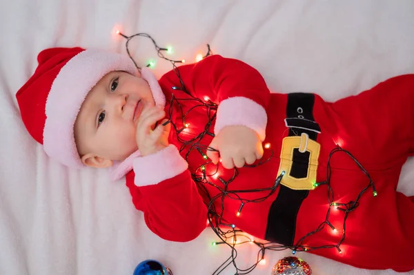 Chłopiec ubrany w strój Mikołaja leży na łóżku i bawi się świąteczną girlandą. — Zdjęcie stockowe